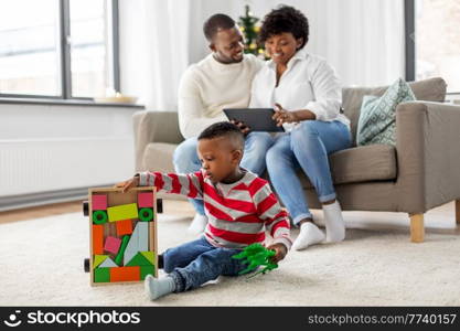 family, winter holidays and people concept - happy african american mother and father using tablet pc computer and baby son playing with toy blocks and dinosaur at home on christmas. happy african american family on christmas at home