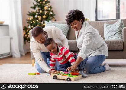 family, winter holidays and people concept - happy african american mother, father and baby son playing with dinosaur and toy blocks at home on christmas. african family with toys on christmas at home