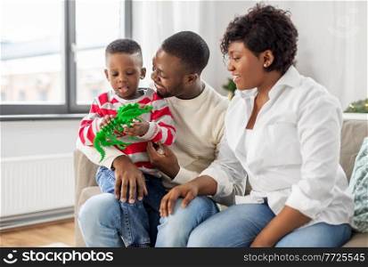 family, winter holidays and people concept - happy african american mother, father and baby son with dinosaur toy at home on christmas. african family with toy on christmas at home