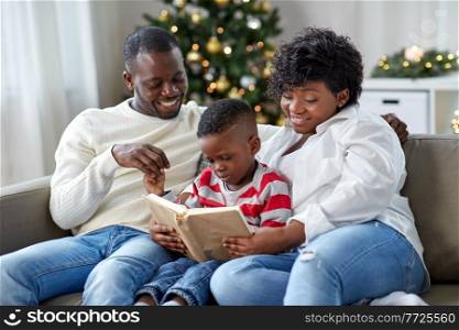 family, winter holidays and people concept - happy african american mother, father and little son reading book at home on christmas. african family reading book on christmas at home