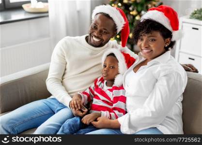 family, winter holidays and people concept - happy african american mother, father and baby son in santa hats at home on christmas. happy african american family on christmas at home