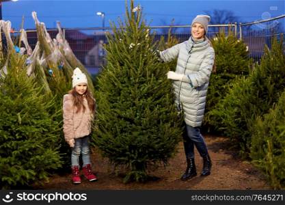 family, winter holidays and people concept - happy mother and little daughter choosing christmas tree at street market in evening. happy family choosing christmas tree at market