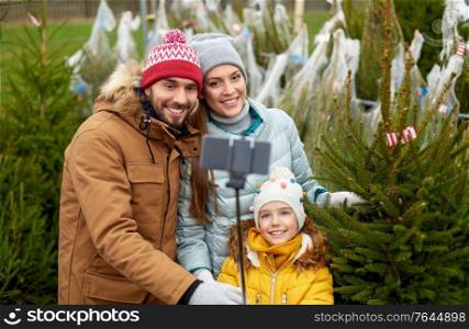 family, winter holidays and people concept - happy mother, father and little daughter buying christmas tree and taking picture with smartphone on selfie stick at street market. family taking selfie with christmas tree at market