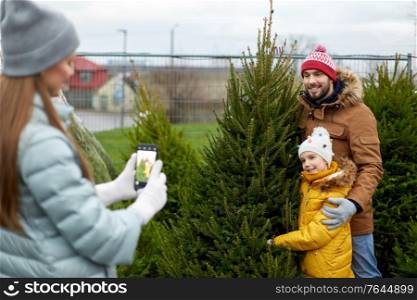 family, winter holidays and people concept - happy mother, father and little daughter buying christmas tree and taking picture with smartphone at street market. family taking picture of christmas tree at market