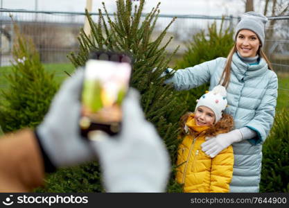 family, winter holidays and people concept - happy mother, father and little daughter buying christmas tree and taking picture with smartphone at street market. family taking picture of christmas tree at market