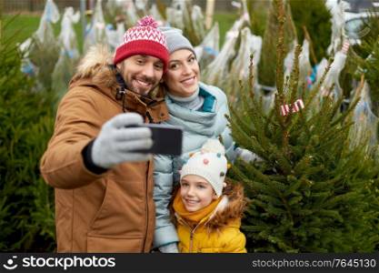 family, winter holidays and people concept - happy mother, father and little daughter buying christmas tree and taking selfie with smartphone at street market. family taking selfie with christmas tree at market