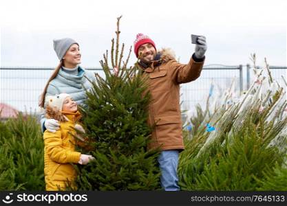 family, winter holidays and people concept - happy mother, father and little daughter buying christmas tree and taking selfie with smartphone at street market. family taking selfie with christmas tree at market