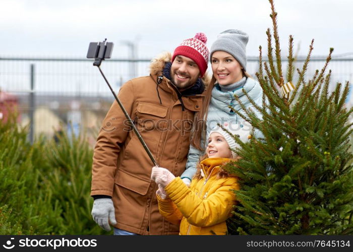 family, winter holidays and people concept - happy mother, father and little daughter buying christmas tree and taking picture with smartphone on selfie stick at street market. family taking selfie with christmas tree at market