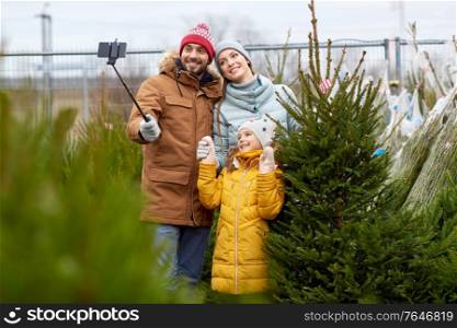 family, winter holidays and people concept - happy mother, father and little daughter buying christmas tree and taking picture with smartphone on selfie stick at street market. family taking selfie with christmas tree at market