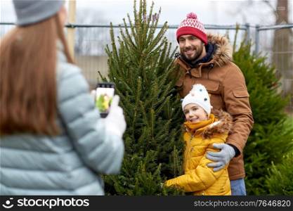 family, winter holidays and people concept - happy mother, father and little daughter buying christmas tree and taking picture with smartphone at street market. family taking picture of christmas tree at market