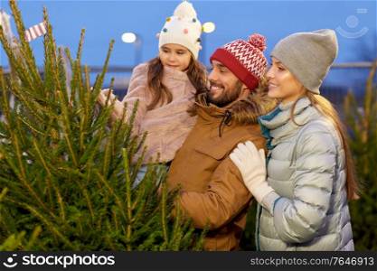 family, winter holidays and people concept - happy mother, father and little daughter choosing christmas tree at street market in evening. happy family choosing christmas tree at market
