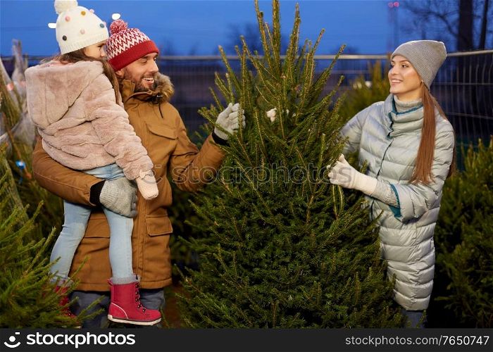 family, winter holidays and people concept - happy mother, father and little daughter choosing christmas tree at street market in evening. happy family choosing christmas tree at market