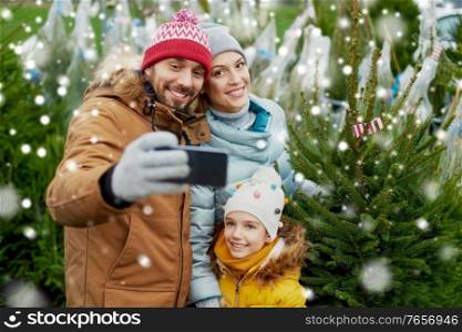 family, winter holidays and people concept - happy mother, father and little daughter buying christmas tree and taking selfie with smartphone at street market over snow. family taking selfie with christmas tree at market