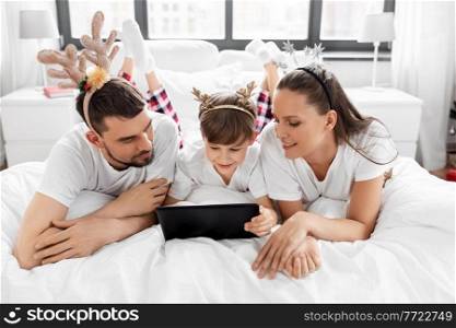 family, winter holidays and people concept - happy mother, father and little son in pajamas with tablet pc computer in bed in christmas morning. family with tablet pc in bed in christmas morning