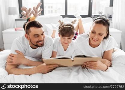 family, winter holidays and people concept - happy mother, father and little son in pajamas reading book in bed in christmas morning. family with book in pajamas in bed on christmas