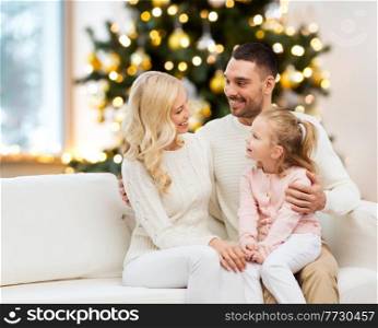 family, winter holidays and people concept - happy mother, father and little daughter sitting on sofa at home over christmas tree background. happy family at home over christmas tree