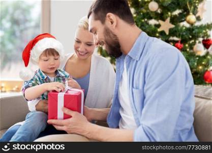 family, winter holidays and people concept - happy mother, father and little son with gift box sitting on sofa at home over christmas tree background. happy family at home with christmas gift box