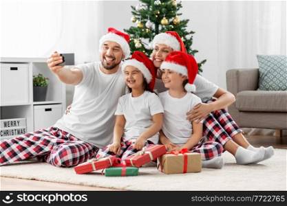 family, winter holidays and people concept - happy mother, father and two daughters in santa hats sitting under christmas tree at home and taking selfie with smartphone. happy family taking selfie on christmas at home