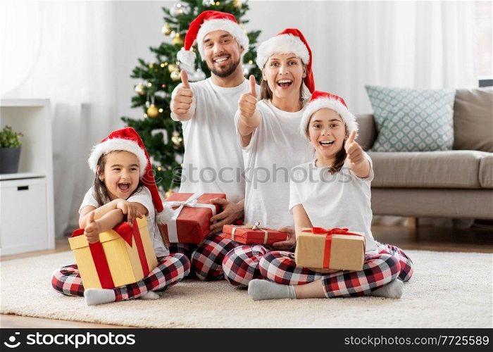 family, winter holidays and people concept - happy mother, father and two daughters in matching pajamas sitting with gifts under christmas tree at home showing thumbs up. happy family sitting under christmas tree at home