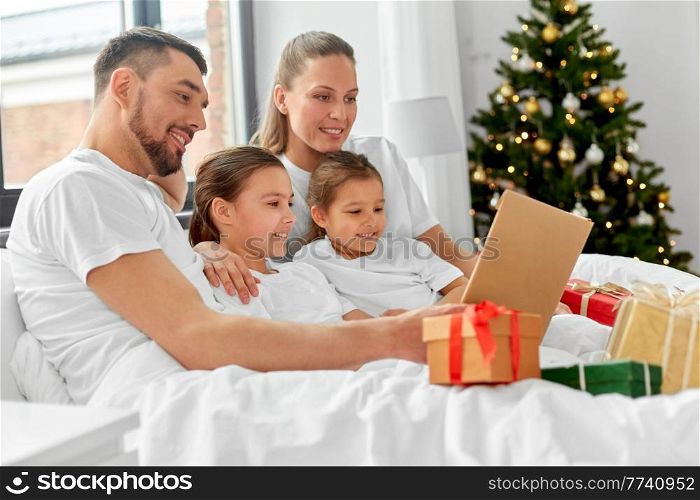 family, winter holidays and people concept - happy mother, father and two daughters in pajamas with christmas gifts reading book in bed at home. happy family reading book in bed on christmas