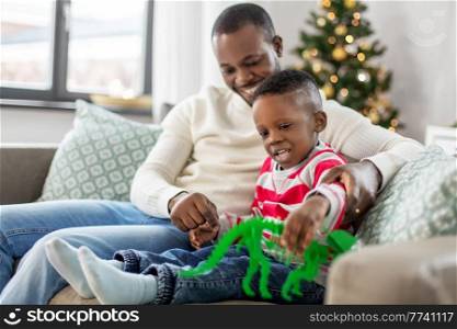 family, winter holidays and people concept - happy smiling african american father and baby son with dinosaur toy at home on christmas. happy father playing with son on christmas at home
