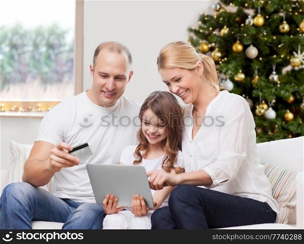 family, winter holidays and technology concept - happy mother, father and daughter with tablet pc computers and credit card shopping online at home over christmas tree lights background. family with tablet pc and credit card on christmas
