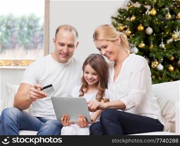 family, winter holidays and technology concept - happy mother, father and daughter with tablet pc computers and credit card shopping online at home over christmas tree lights background. family with tablet pc and credit card on christmas