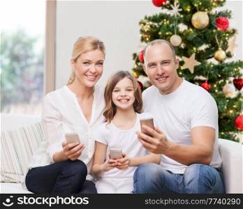 family, winter holidays and technology concept - happy mother, father and daughter with smartphones at home over christmas tree lights background. happy family with smartphones at home on christmas