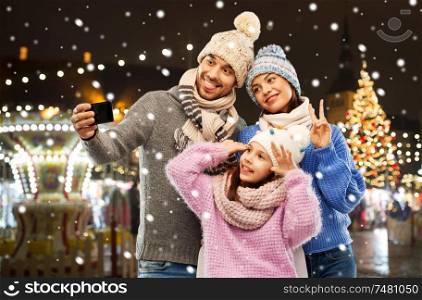 family, winter holidays and technology concept - happy mother, father and little daughter in winter clothes taking selfie by smartphone over christmas market in old town of tallinn city background. happy family taking selfie at christmas market
