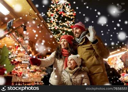 family, winter holidays and technology concept - happy mother, father and little daughter taking picture by smartphone on selfie stick at christmas market in tallinn, estonia over snow. happy family taking selfie at christmas market