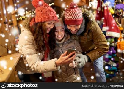 family, winter holidays and technology concept - happy mother, father and little daughter with smartphone at christmas market in evening over snow. happy family with smartphone at christmas market