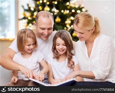 family, winter holidays and technology concept - happy mother, father and two daughters sitting on sofa and reading book at home over christmas tree lights background. happy family reading book at home on christmas