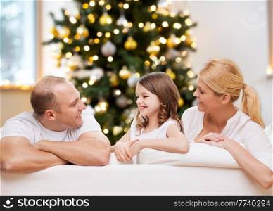 family, winter holidays and technology concept - portrait of happy mother, father and daughter sitting on sofa at home over christmas tree lights background. portrait of happy family at home on christmas