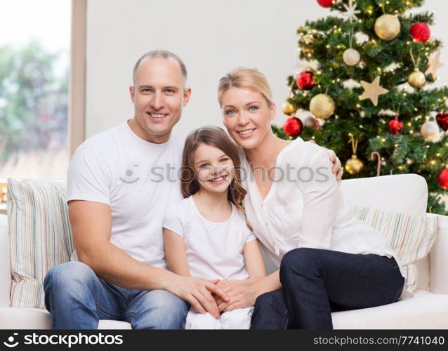 family, winter holidays and technology concept - portrait of happy mother, father and daughter sitting on sofa at home over christmas tree lights background. portrait of happy family at home on christmas