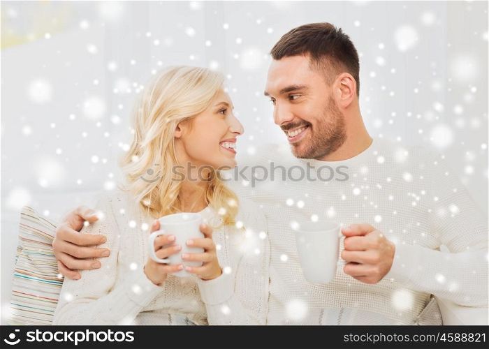 family, winter, holidays, love and people concept - happy couple covered by plaid with cups drinking tea and sitting on sofa at home