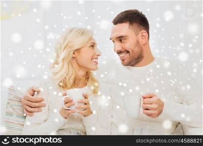 family, winter, holidays, love and people concept - happy couple covered by plaid with cups drinking tea and sitting on sofa at home