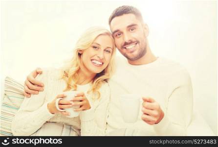 family, winter, holidays, love and people concept - happy couple covered by plaid with cups drinking tea and sitting on sofa at home. happy couple with cups drinking tea at home