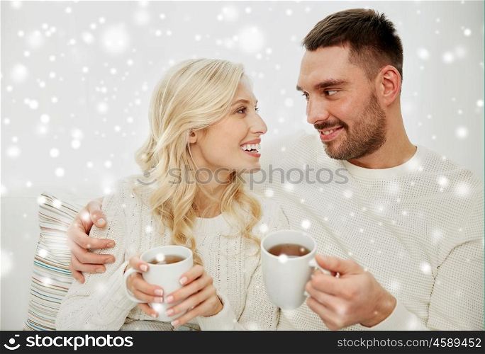 family, winter, holidays, love and people concept - happy couple with cups drinking tea and sitting on sofa at home