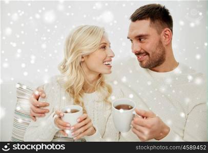 family, winter, holidays, love and people concept - happy couple with cups drinking tea and sitting on sofa at home