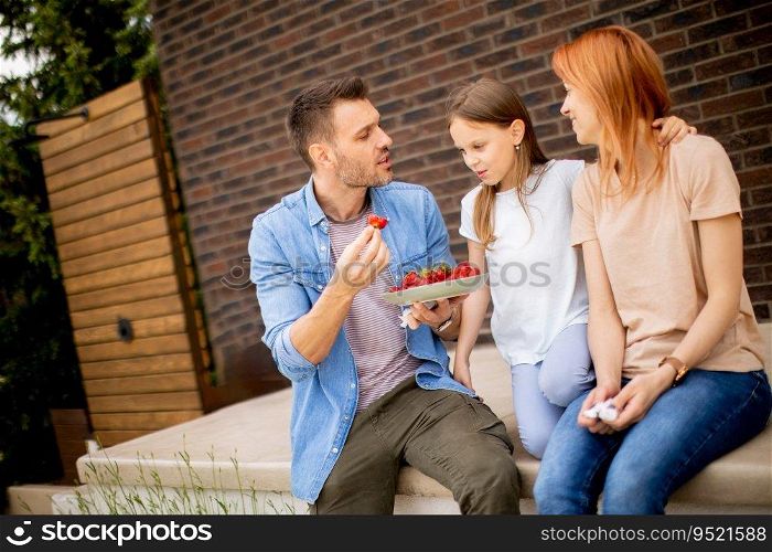 Family with a mother, father and daughter sitting outside on a steps of a front porch of a brick house and eating strawberries