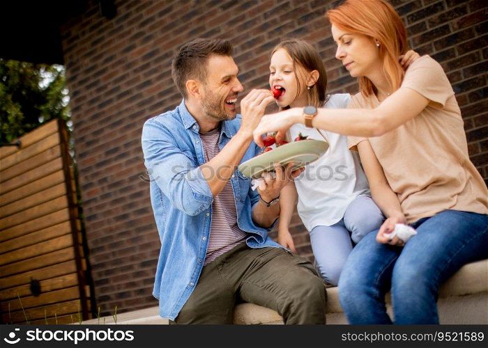 Family with a mother, father and daughter sitting outside on a steps of a front porch of a brick house and eating strawberries
