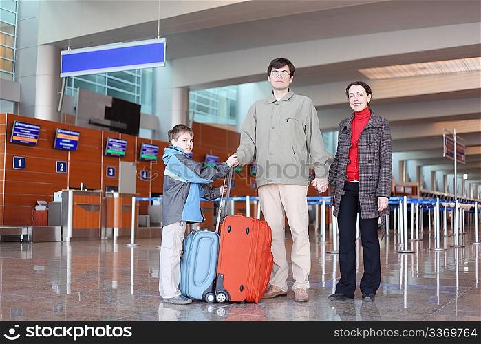 family with boy standing in airport hall with suitcases full body looking at camera