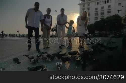 Family with child is going to feed pigeons when little boy walking through the flock of bird. Aristotelous Square at sunset