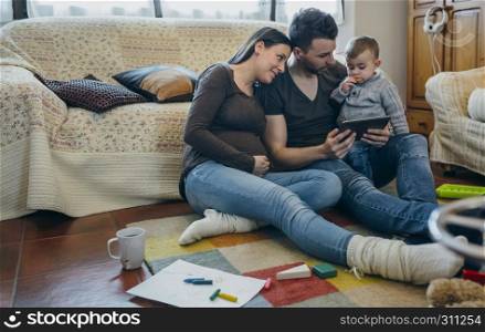 Family with small child and pregnant mother looking at the tablet sitting on the carpet. Family with child and pregnant mother looking tablet