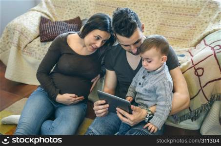 Family with small child and pregnant mother looking at the tablet sitting on the carpet. Family with child and pregnant mother looking tablet