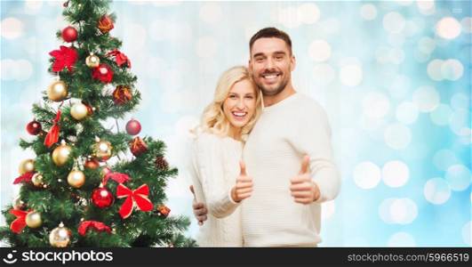 family, x-mas, winter holidays and people concept - happy couple showing thumbs up with christmas tree at home over blue lights background