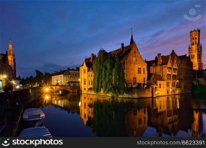 Famous view of Bruges tourist landmark attraction - Rozenhoedkaai canal with Belfry and old houses along canal with tree in the night. Brugge, Belgium. Famous view of Bruges, Belgium
