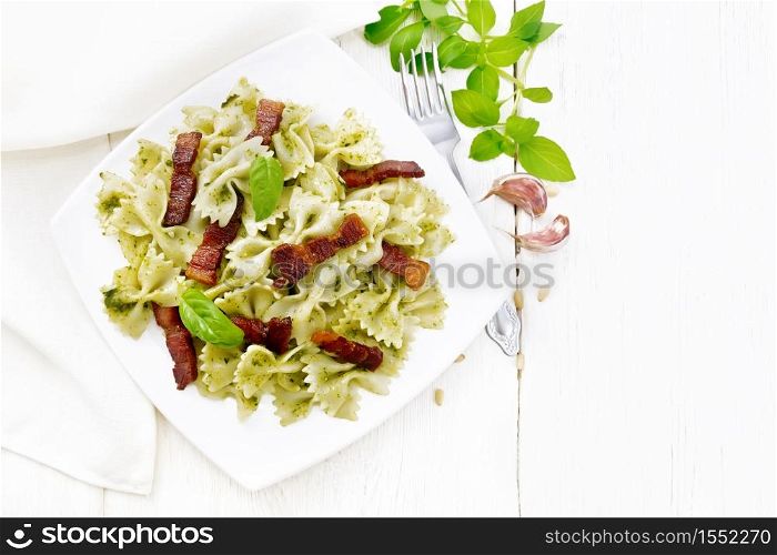 Farfalle pasta with pesto sauce, fried bacon and basil in a plate, garlic, fork and napkin on wooden board background from above