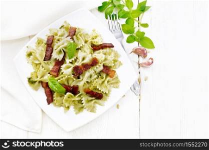 Farfalle pasta with pesto sauce, fried bacon and basil in a plate, garlic, fork and napkin on wooden board background from above