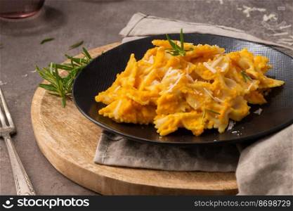 Farfalle pasta with pumpkin sauce and parmesan cheese decorated with rosemary on a black ceramic plate. Creamy cheesy vegan pumpkin pasta. Kitchen counter top.